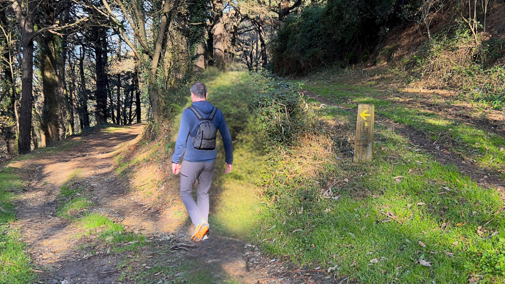 Pilgrim walks on the Camino de Santiago. He is wearing The Mighty Mini Unisex Backpack from Ultreiatrek. He has just passed a mile marker and opted for the lower trail as the yellow arrow indicates.