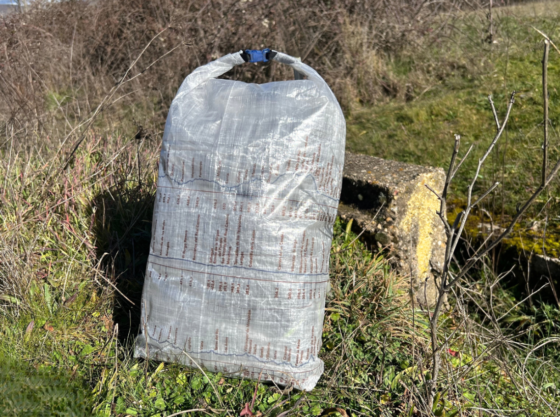 Waterproof bag with Ultreiatrek design with all the towns and kilometers on the Camino Francés. Clear fabric with route in blue and towns in brown are visible on the bag. Blue buckle attaches on top. Bag sits on the grass on the Camino de Santiago trail near a yellow arrow.