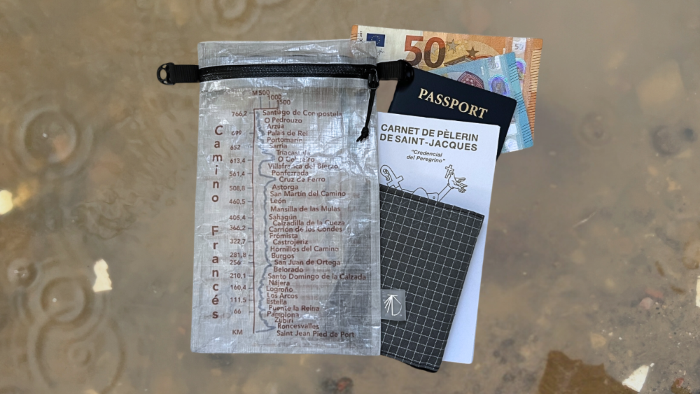 Ultreiatrek's Dyneema® Valuables Holder Bag with the elevation gain and kilometers from Saint Jean Pied de Port to Santiago de Compostela. It is featured on a frosted background and shows items that fit in the holder such as a wallet, pilgrim's credential, passport and money.